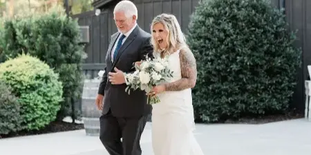 A bride and her father walking in front of a dark wooden barn with string lights.