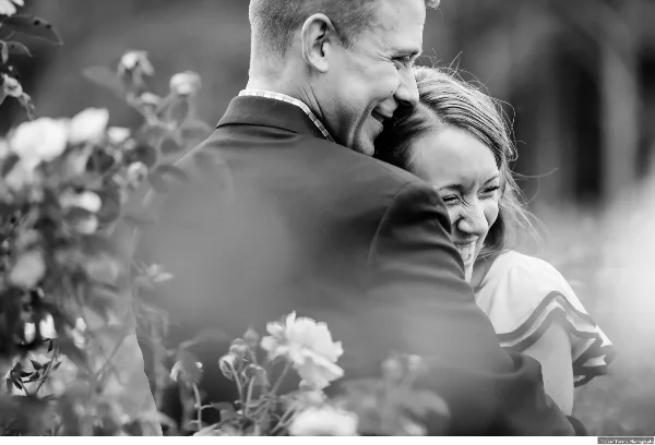 A black and white photograph of a smiling couple embracing in a garden with flowers in the foreground.