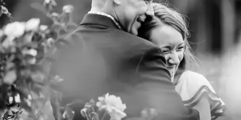 A black and white photograph of a smiling couple embracing in a garden with flowers in the foreground.