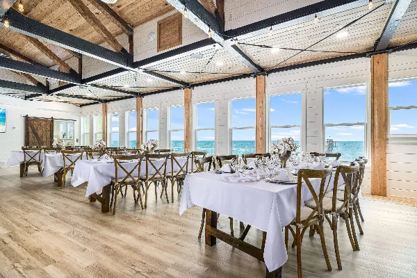 A spacious, light-filled wedding reception hall with white tablecloths, wooden chairs, and large windows overlooking the ocean.