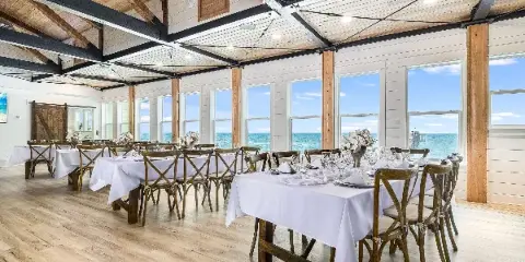 A spacious, light-filled wedding reception hall with white tablecloths, wooden chairs, and large windows overlooking the ocean.