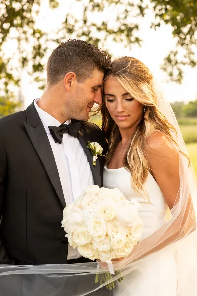 A close-up portrait of a bride and groom in wedding attire, with the bride holding a white rose bouquet.