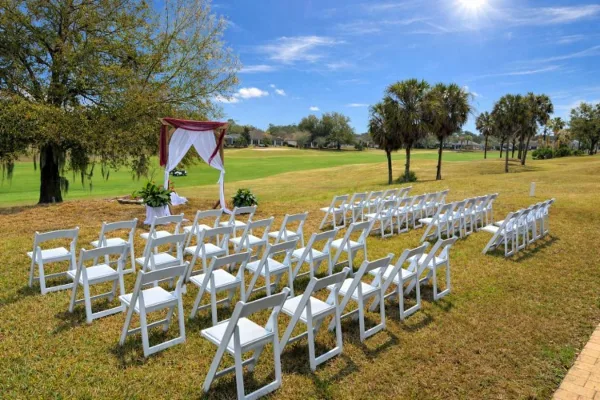 Outdoor wedding ceremony setup with rows of white chairs and a decorated arch on a grassy field.