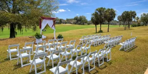 Outdoor wedding ceremony setup with rows of white chairs and a decorated arch on a grassy field.