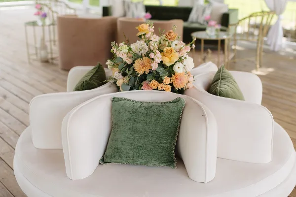 A lush floral arrangement featuring yellow roses and pink blooms placed on a white curved sofa with green velvet pillows in a bright event venue.