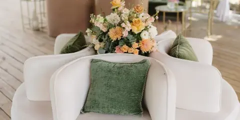 A lush floral arrangement featuring yellow roses and pink blooms placed on a white curved sofa with green velvet pillows in a bright event venue.