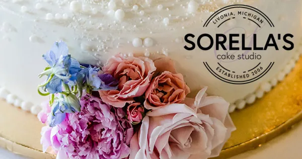 A close-up of a white wedding cake decorated with pink and purple fresh flowers, featuring the Sorella's Cake Studio logo.