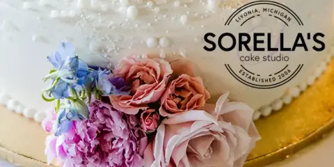 A close-up of a white wedding cake decorated with pink and purple fresh flowers, featuring the Sorella's Cake Studio logo.