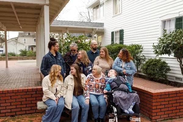 A multi-generational family posing together on a brick porch outside a white house, including a child in a wheelchair.