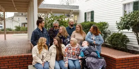A multi-generational family posing together on a brick porch outside a white house, including a child in a wheelchair.