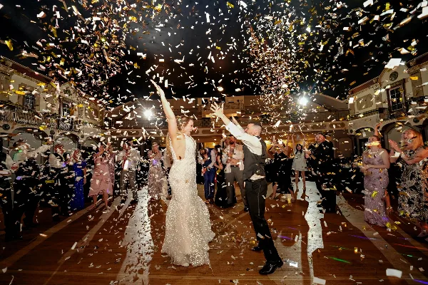 A bride and groom dancing on a wooden floor while a large amount of gold and white confetti falls from the ceiling, surrounded by guests.