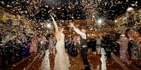 A bride and groom dancing on a wooden floor while a large amount of gold and white confetti falls from the ceiling, surrounded by guests.