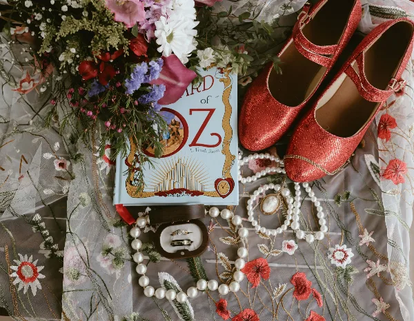 A flat lay photograph of a wedding dress, red sparkly shoes, a bouquet of flowers, a book, and wedding rings.