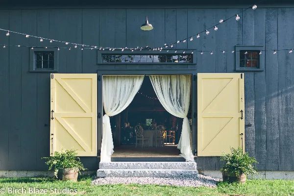 Exterior view of a rustic barn wedding venue featuring dark grey siding, large yellow barn doors, and string lights.