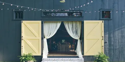 Exterior view of a rustic barn wedding venue featuring dark grey siding, large yellow barn doors, and string lights.