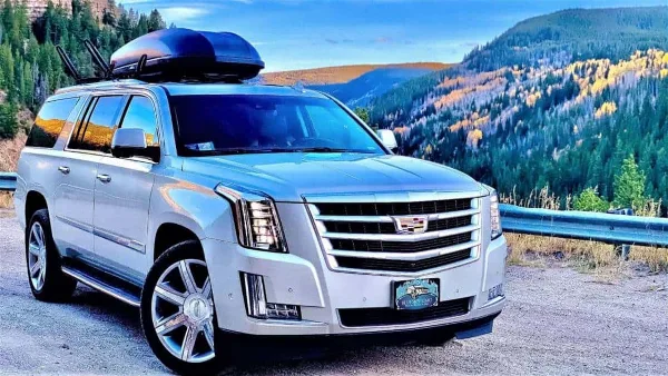 A silver luxury SUV with a Blue Sky Limo license plate frame parked on a mountain road with forested hills in the background.