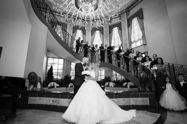 A black and white photograph of a bride and groom standing in a grand ballroom with a large chandelier and a curved staircase.