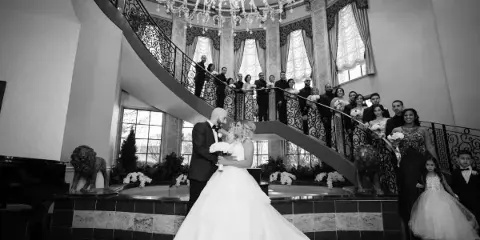 A black and white photograph of a bride and groom standing in a grand ballroom with a large chandelier and a curved staircase.