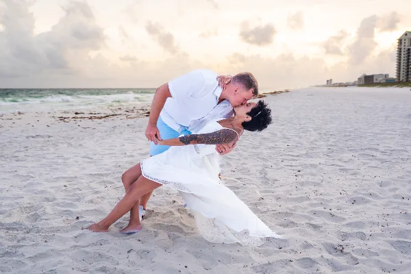 A bride and groom sharing a kiss on a sandy beach at sunset.