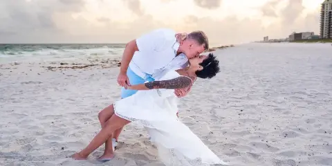A bride and groom sharing a kiss on a sandy beach at sunset.