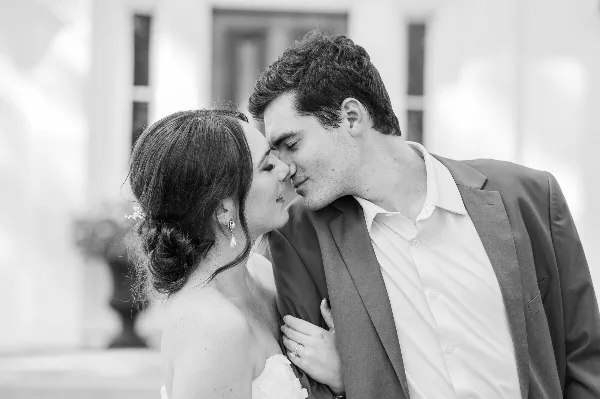 Black and white photograph of a bride and groom sharing a tender moment with foreheads touching in front of a building.