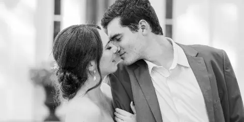 Black and white photograph of a bride and groom sharing a tender moment with foreheads touching in front of a building.