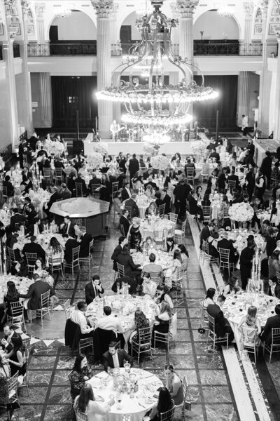 A black and white photograph of a crowded wedding reception hall with guests seated at round tables under a large chandelier.