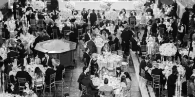 A black and white photograph of a crowded wedding reception hall with guests seated at round tables under a large chandelier.