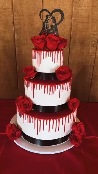A three-tiered white wedding cake decorated with red drip icing, red roses, and a silhouette topper of a couple.