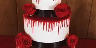 A three-tiered white wedding cake decorated with red drip icing, red roses, and a silhouette topper of a couple.