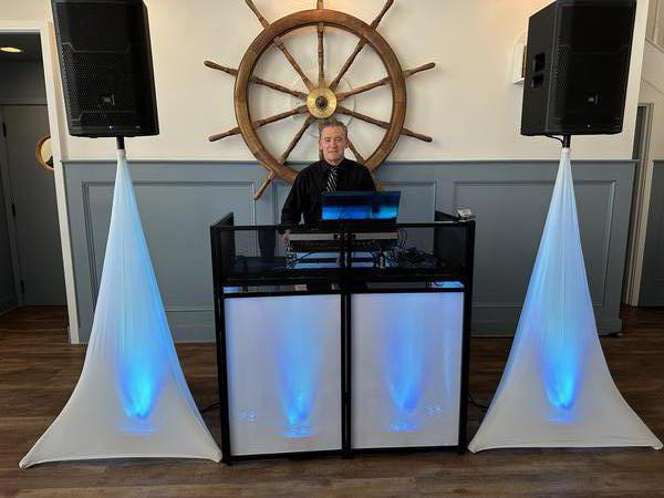 A DJ performing at a booth with professional audio equipment and uplighting, set against a wall with a large ship wheel decoration.