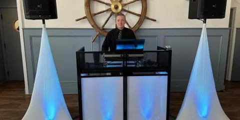 A DJ performing at a booth with professional audio equipment and uplighting, set against a wall with a large ship wheel decoration.