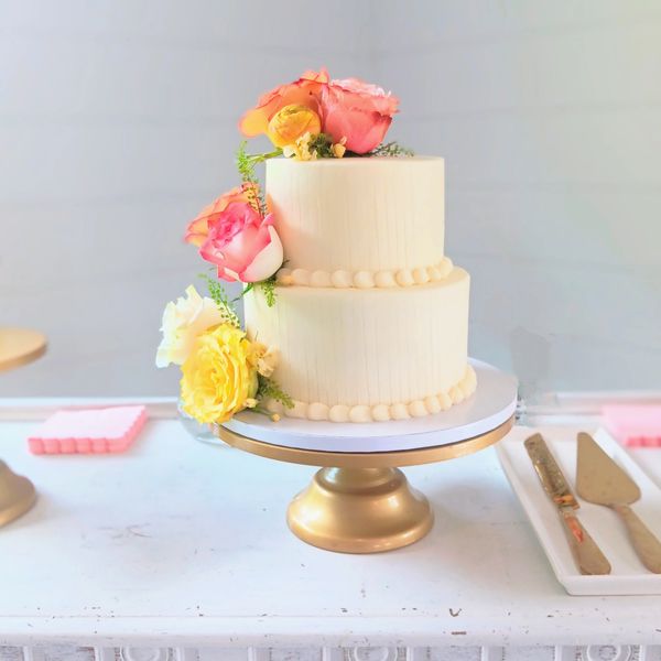 A two-tier white wedding cake decorated with pink and yellow roses on a gold stand.