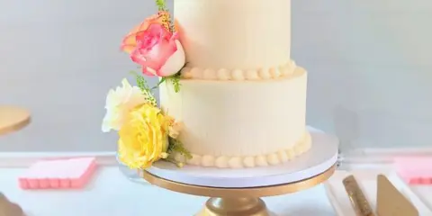 A two-tier white wedding cake decorated with pink and yellow roses on a gold stand.