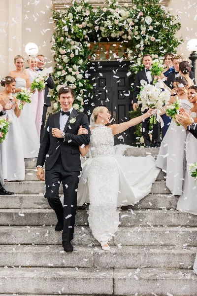 A bride and groom walking down stone steps with their wedding party while confetti falls around them.