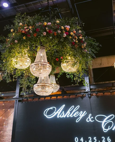 A suspended floral installation with chandeliers and a neon sign reading 'Ashley & C' at a wedding venue.