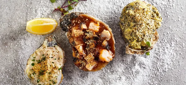 Three prepared oysters with various toppings and a lemon wedge on a textured surface.