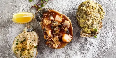Three prepared oysters with various toppings and a lemon wedge on a textured surface.