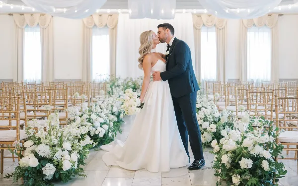A bride and groom kissing in a wedding ceremony aisle lined with white floral arrangements and bamboo chairs.