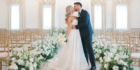 A bride and groom kissing in a wedding ceremony aisle lined with white floral arrangements and bamboo chairs.