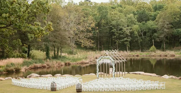 An outdoor wedding ceremony setup with white chairs and a floral arch by a pond.