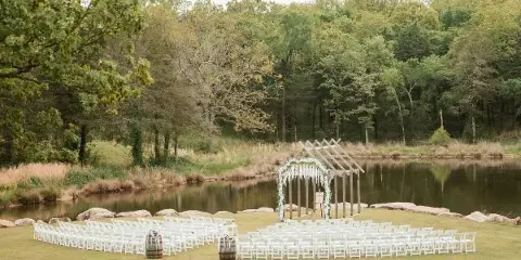 An outdoor wedding ceremony setup with white chairs and a floral arch by a pond.