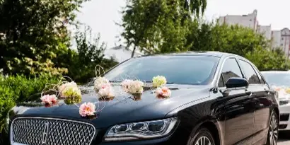 A black luxury sedan decorated with pink and white flowers on the hood, parked outdoors.