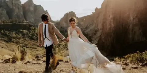 A bride and groom holding hands while walking through a rocky, mountainous landscape.