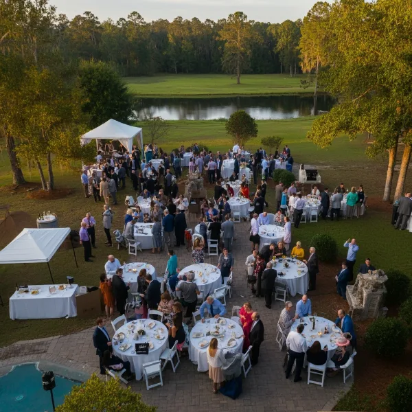 An aerial view of an outdoor wedding reception with white tents, round tables, and white chairs set up on a lawn near a pond.