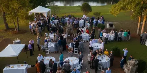 An aerial view of an outdoor wedding reception with white tents, round tables, and white chairs set up on a lawn near a pond.