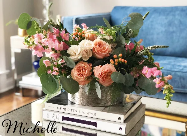 A lush floral arrangement featuring peach roses, pink stock, and eucalyptus in a metallic container, displayed on a stack of books in a styled interior setting.