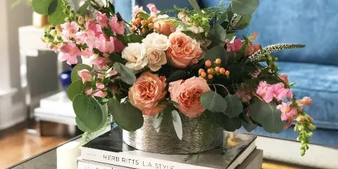 A lush floral arrangement featuring peach roses, pink stock, and eucalyptus in a metallic container, displayed on a stack of books in a styled interior setting.