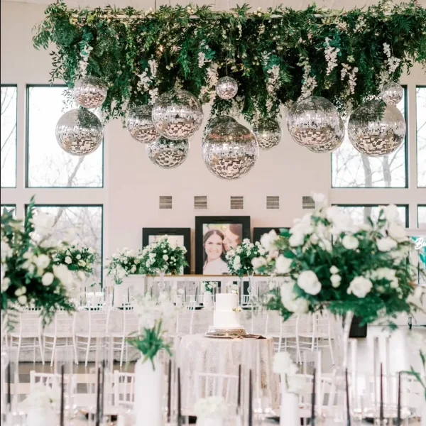 A wedding reception venue decorated with a large overhead floral installation, hanging disco balls, and white floral centerpieces on tables.