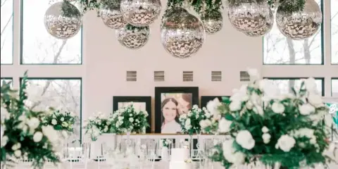 A wedding reception venue decorated with a large overhead floral installation, hanging disco balls, and white floral centerpieces on tables.